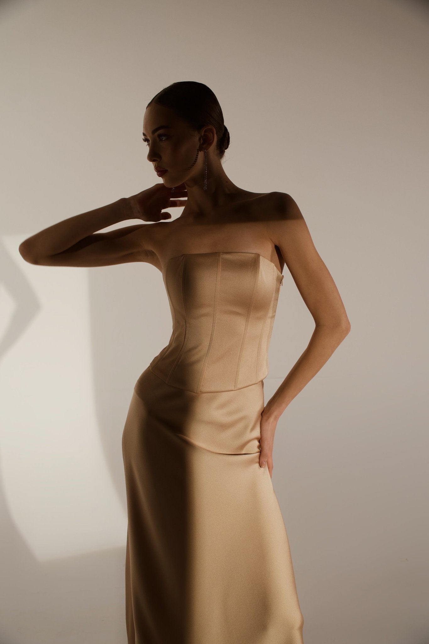 A woman in a strapless, form-fitting beige dress strikes an elegant pose. The lighting casts a dramatic shadow on the wall behind her, accentuating the contours of her Regina corset top and creating a striking contrast between light and dark areas.