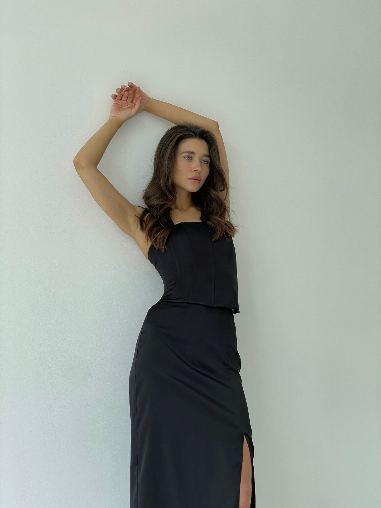 A woman with long brown hair is posing against a plain white background. She is wearing the Regina corset top, which features sculpting satin fabric and a sleeveless design. Her arms are raised above her head, with one hand grasping the other arm's elbow, and she is looking slightly to the side.