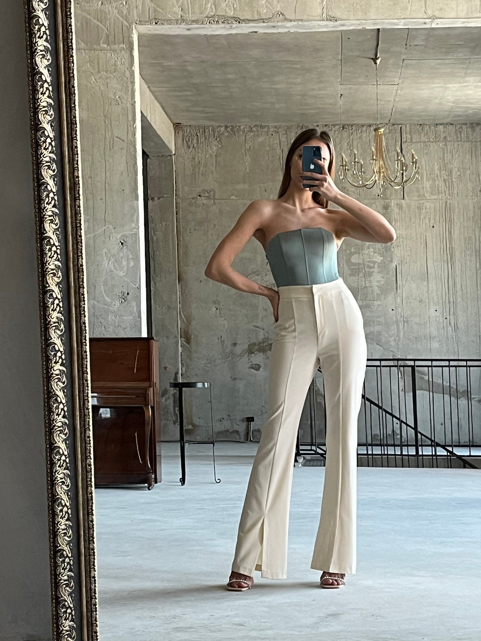A woman stands in a spacious, industrial-style room, taking a mirror selfie. She is wearing the Regina corset top paired with high-waisted white trousers and heels. A chandelier hangs from the ceiling and a wooden piano is visible in the background.