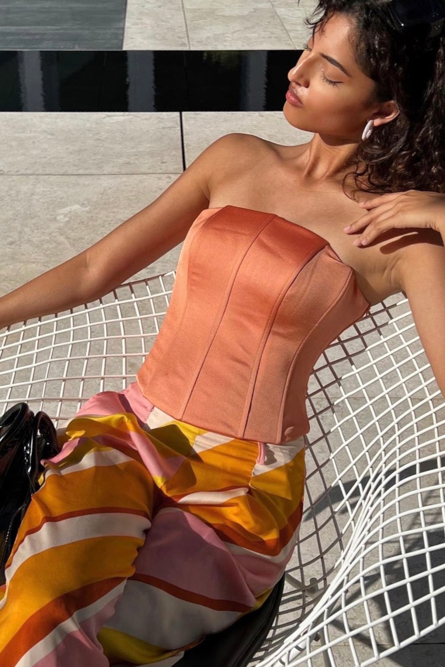 A woman with curly hair lounges on a white wire chair outdoors, dressed in a Regina corset top in peach and colorful striped pants. Sunlight casts shadows, highlighting her relaxed and serene expression. A black handbag is placed beside her, completing the Bridgerton-esque look beautifully.