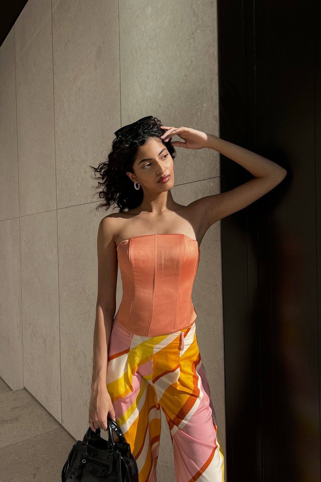 A woman with curly hair stands against a wall, wearing a strapless orange Regina corset top and brightly patterned pants in shades of pink, yellow, and white. She holds a small black handbag and shades her eyes with one hand. The background is a mix of concrete and dark paneling.