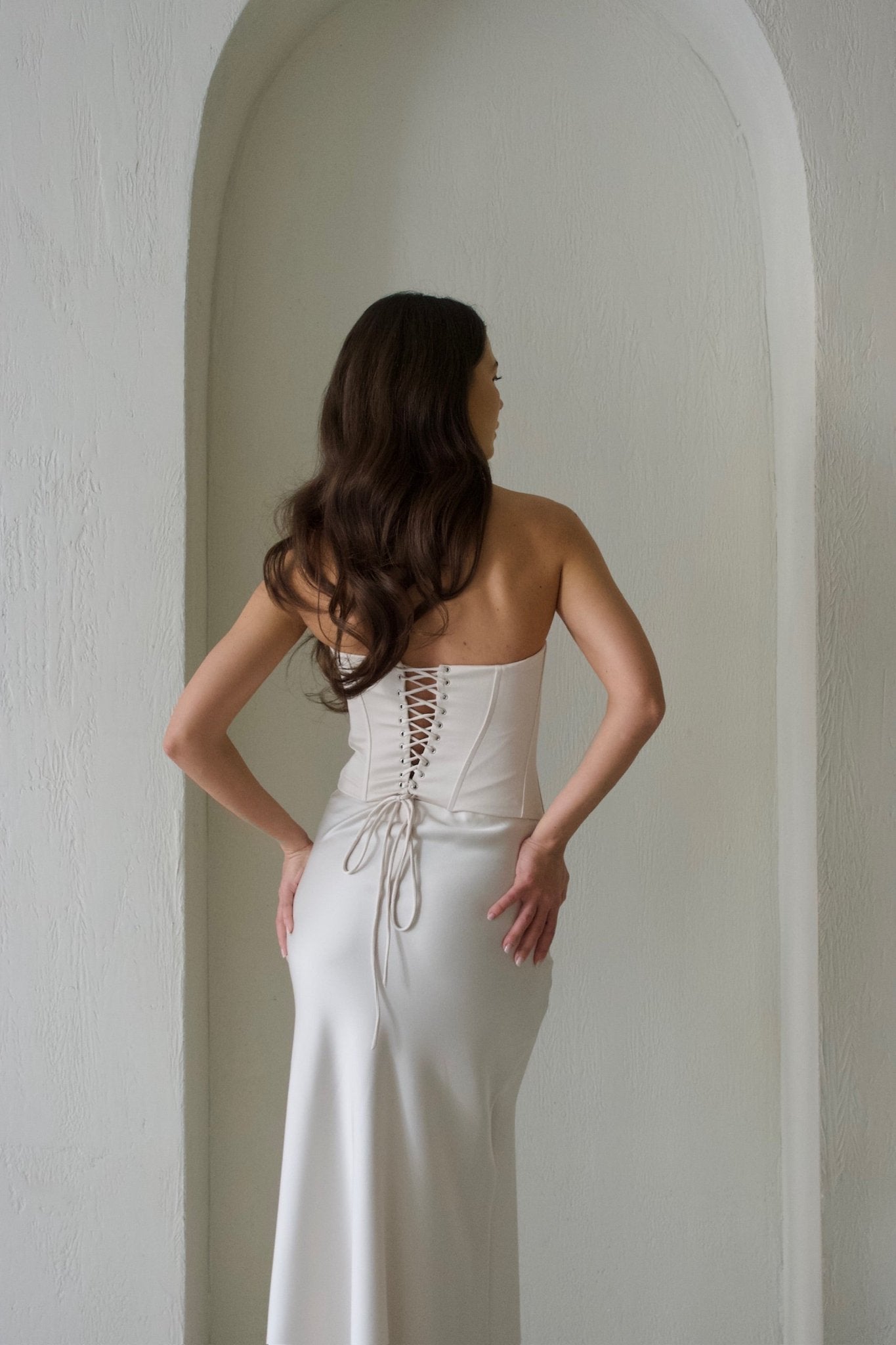 A woman stands with her back to the camera, elegantly dressed in the Regina Corset Set featuring a strapless, form-fitting white dress. The ensemble showcases a lace-up corset back and a long, flowing maxi skirt made from luxurious satin fabric. Her long, wavy brown hair cascades down as she poses against a light-colored, textured archway backdrop. Pre-order now for the exclusive Regina Corset Set with Maxi Skirt.
