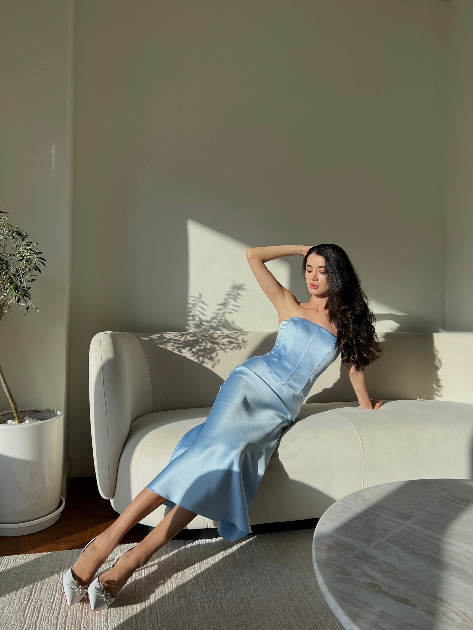A woman in a Regina corset top, which has a striking resemblance to a Bridgerton-inspired blue strapless dress, reclines on a cream-colored sofa in a softly lit room. She wears white shoes and her long dark hair cascades over her shoulders. A potted plant and a round marble table are visible. Sunlight casts shadows on the wall.