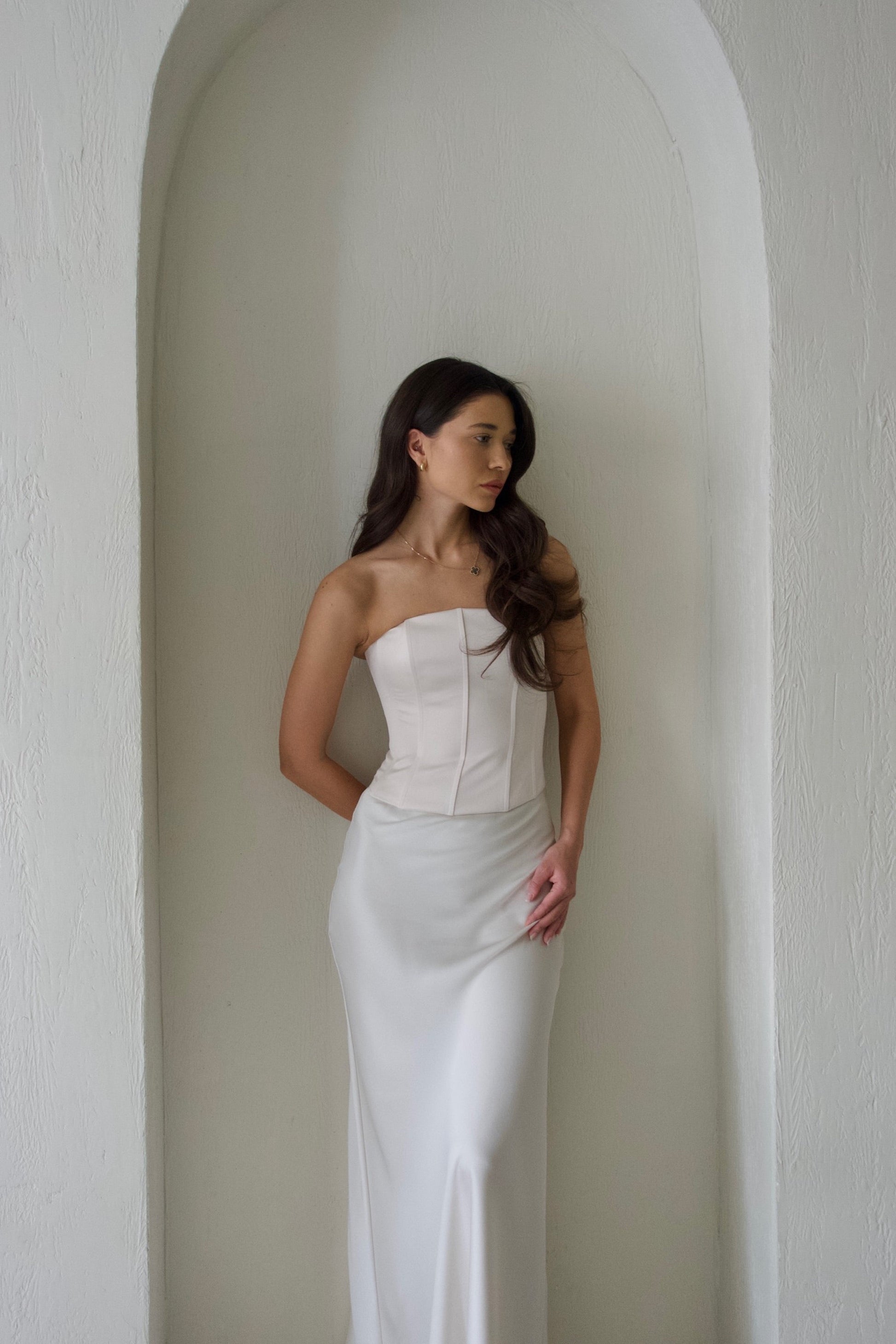 A woman wearing the Regina Corset Set in White leans against a rounded alcove with textured walls. Her long, dark hair cascades over her shoulder as she gazes to the side. The setting is minimal and elegant.