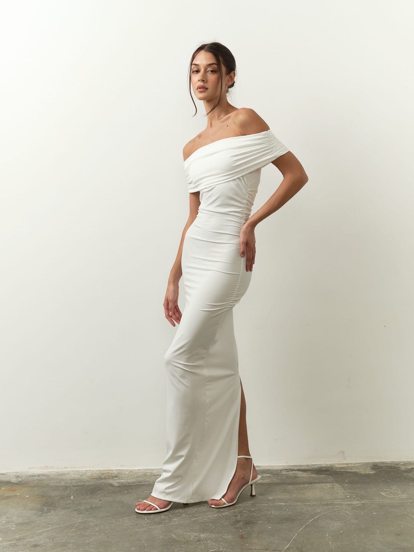 A woman poses in the Scarlett Dress, showcasing its off-the-shoulder design and feminine silhouette. The dress features a form-fitting style with a side slit. She complements her look with high-heeled sandals and an elegant updo. The setting includes a plain white wall and a concrete floor backdrop.