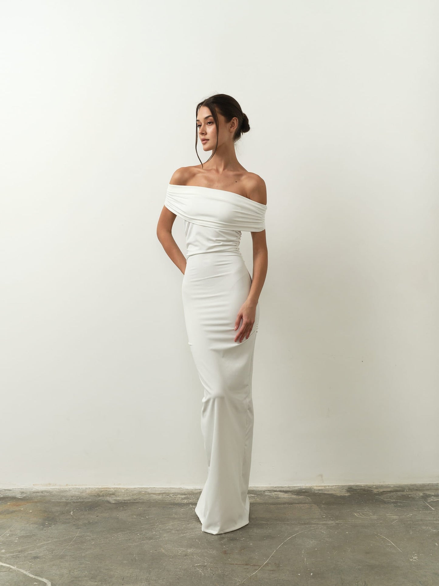 A woman in the elegant Scarlett Dress, an off-the-shoulder white gown, poses against a plain white background. The fitted dress beautifully highlights her feminine silhouette, emphasizing a minimalist and sophisticated style. Her hair is styled in an updo.