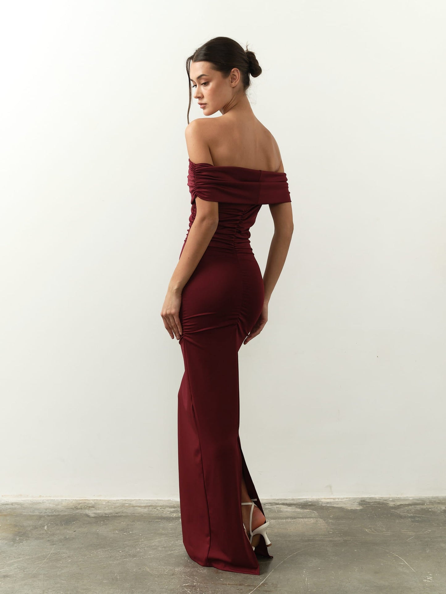 A woman in a fitted Scarlett Dress, an off-the-shoulder burgundy gown, embraces her feminine silhouette as she stands on a bare concrete floor against a plain white wall. Her gaze is downward, and her hair is styled in a neat bun.