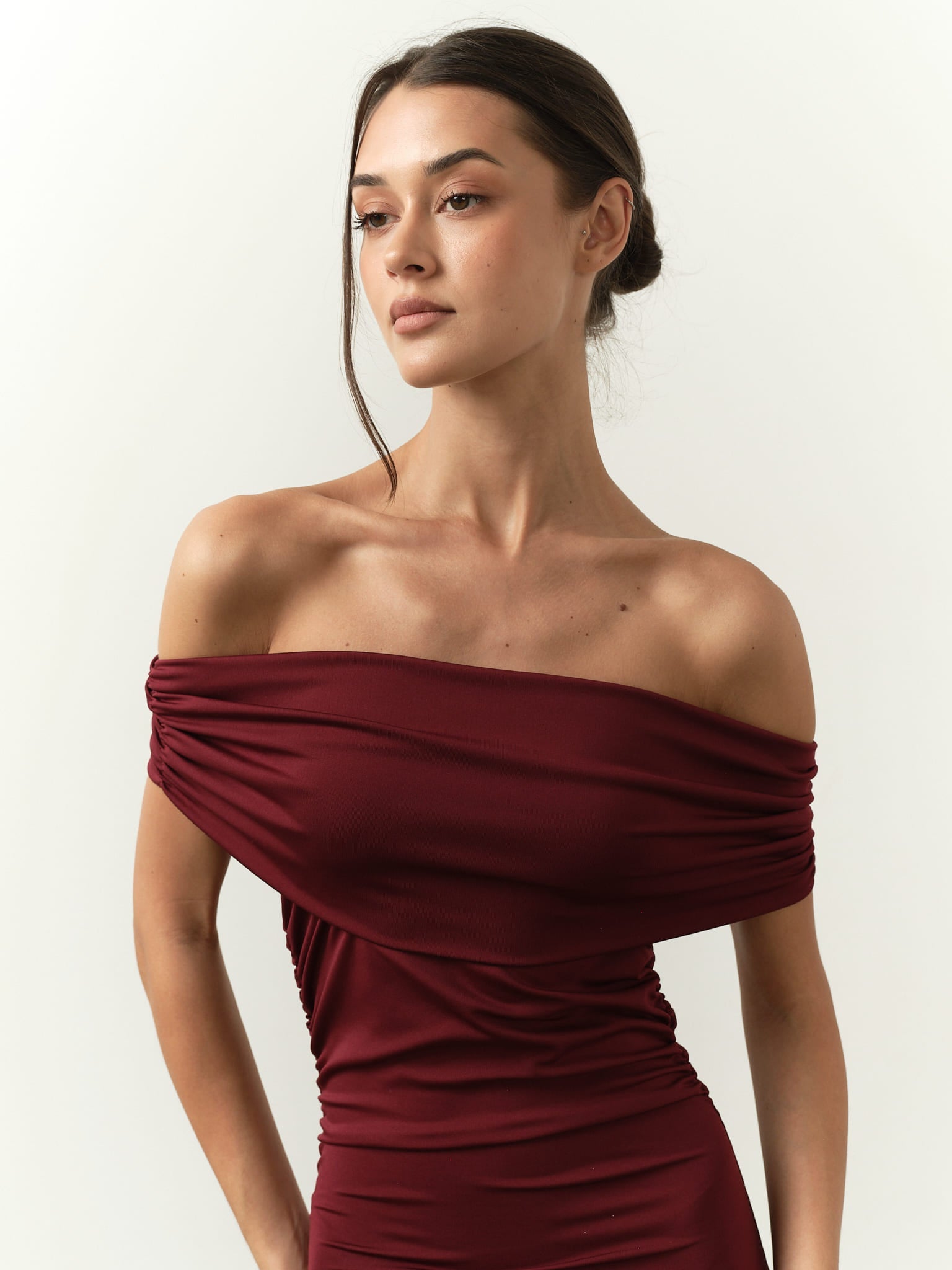 A person with long, dark hair tied back is wearing the Scarlett Dress, an off-the-shoulder burgundy piece that accentuates a feminine silhouette. The background is plain, and the person gazes slightly to the side with a composed expression.