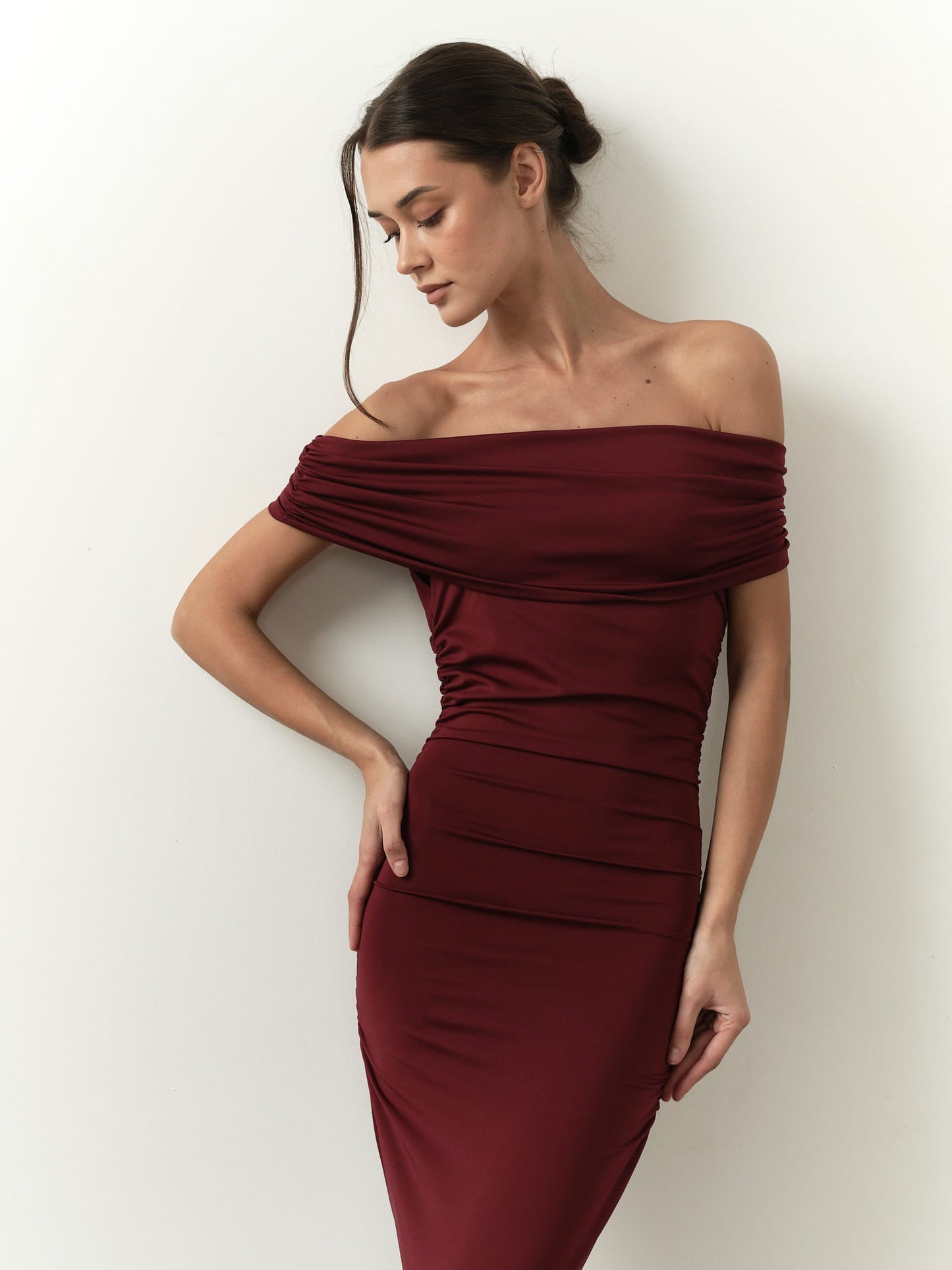 A woman in the Scarlett Dress, an off-the-shoulder burgundy gown, poses against a plain background, accentuating her feminine silhouette. Her hair is styled in an elegant updo as she gazes down with a serene expression.