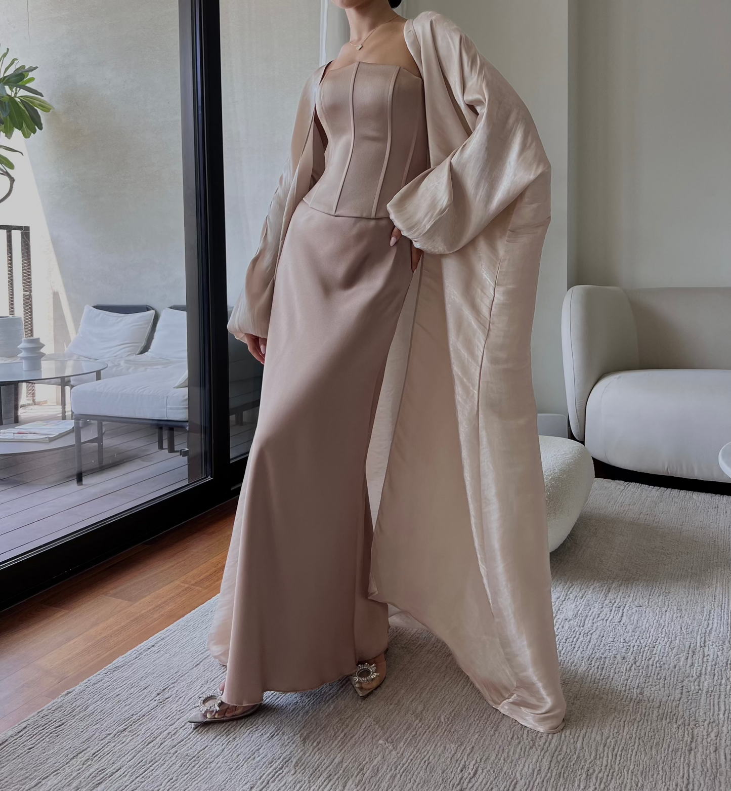 A person is standing indoors wearing a sophisticated light pink floor-length gown with a corset bodice. Complemented by a matching, draped overcoat, they pose with one hand on their hip. The room features modern, minimalist furniture and large windows. Consider pairing it with our Silky maxi skirt for added elegance.