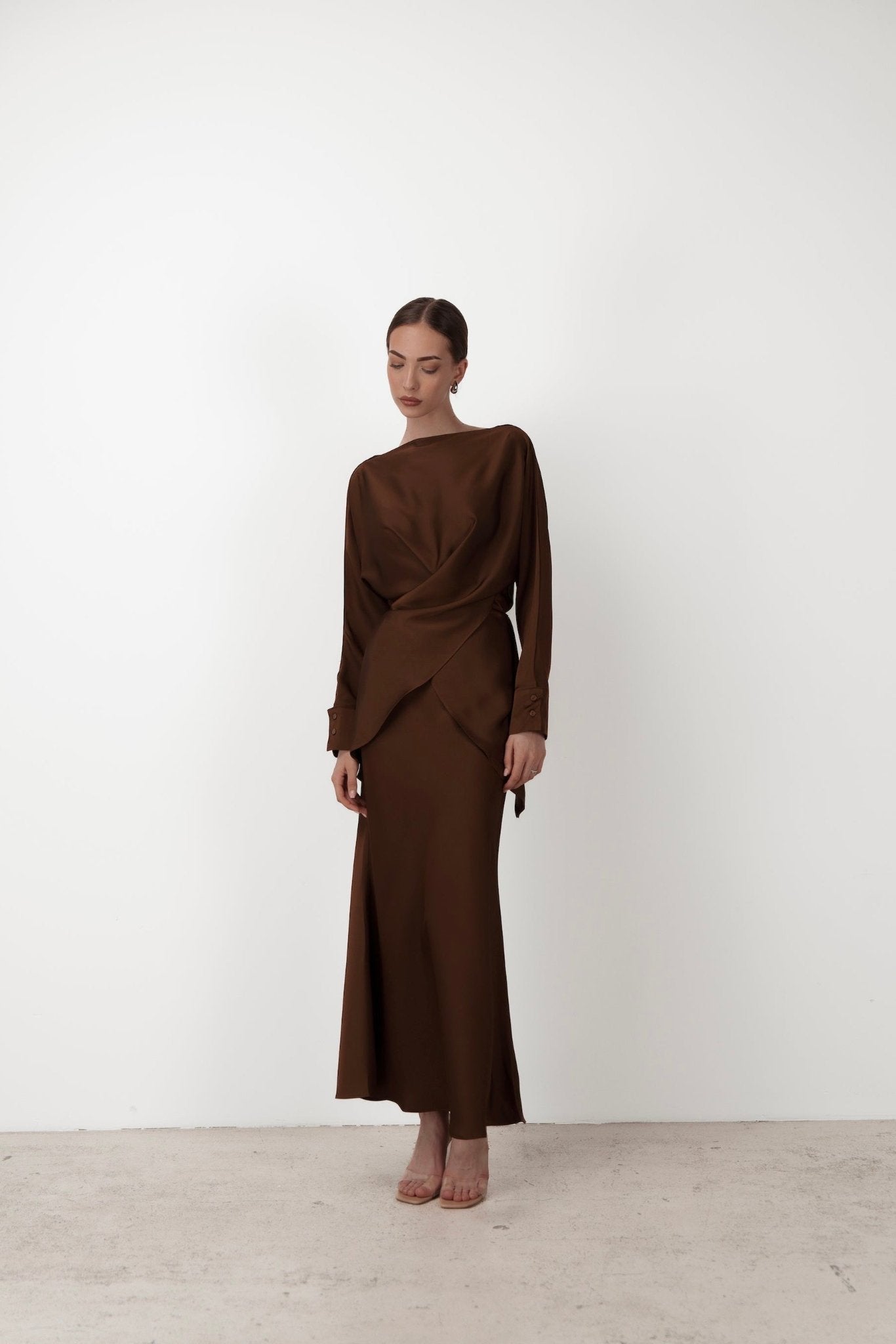 A woman stands against a plain white background, wearing the Tessa set—a long, chocolate-brown dress featuring elegant balloon sleeves and a subtle draped design on the front. The luxurious silky satin fabric enhances the overall look. She has her hair styled in a low bun and is looking slightly downward, paired with nude-colored heels.