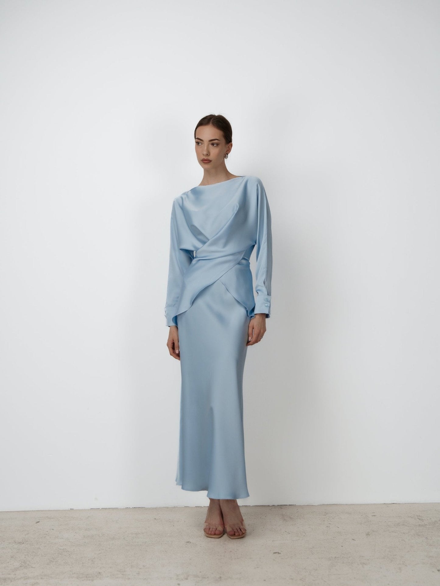 A woman stands against a white background, wearing the elegant Tessa set—a flowing, light blue long-sleeved dress with draped detail across the waist. She has her hair pulled back and is posing with a neutral expression, hands resting gently in front of her. The luxurious ensemble is completed by nude heels, exuding modern sophistication.